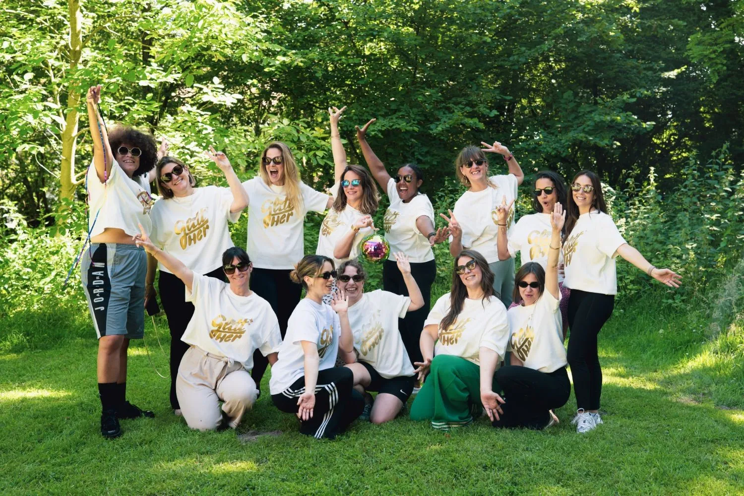 Les participantes de l'Échappée de juin posent ensemble dans l'herbe, en t-shirts Glitt'Her et lunettes de soleil, autour d'une boule à facettes